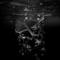 A dark metallic four-leaf clover pendant with a small hanging loop, floating underwater amidst rising bubbles against a black background.