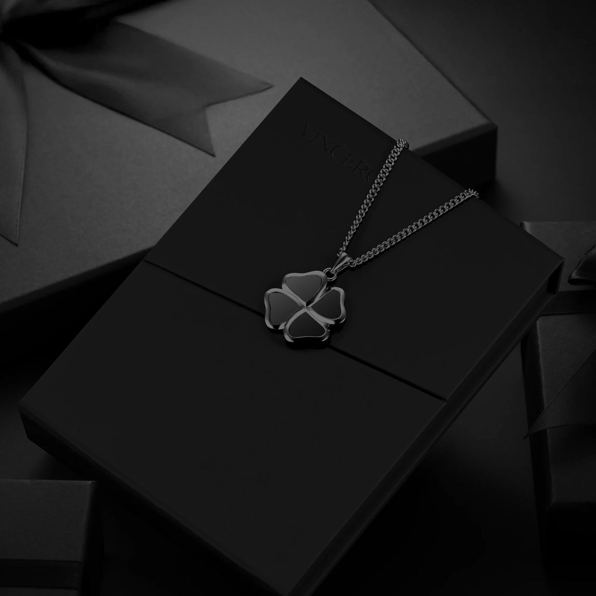 A silver-toned clover pendant necklace displayed on a black presentation box, with dark gift packaging in the background.