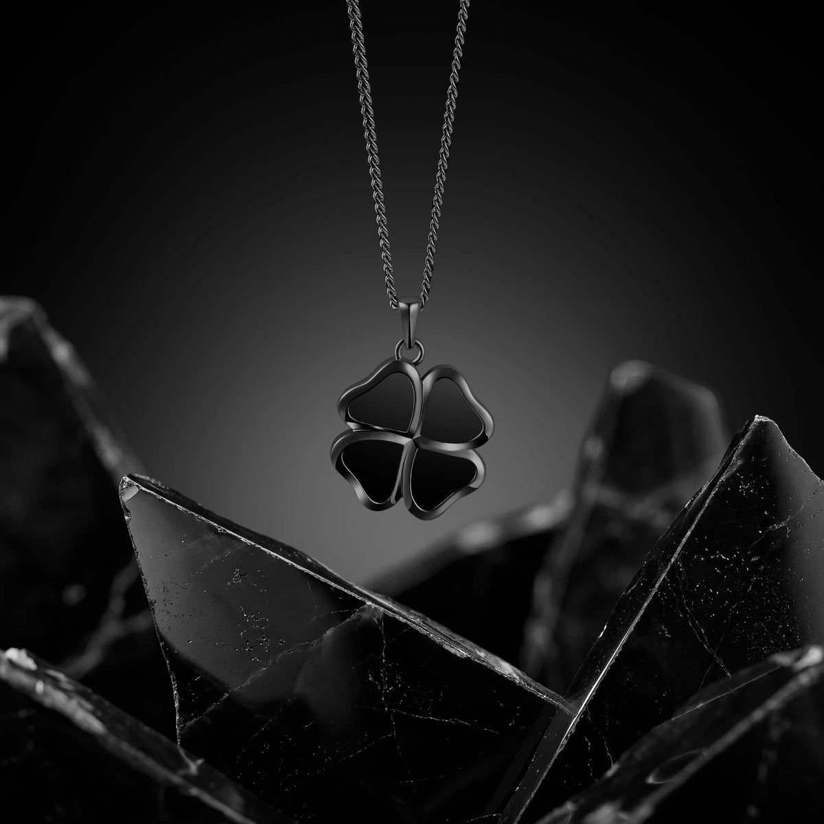 A black four-leaf clover pendant necklace on a silver chain, displayed against a dark gray gradient background with sharp black crystal-like props in the foreground.