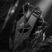 A black metal four-leaf clover pendant necklace on a matching chain, displayed on a faceted black crystal against a dark, moody background.