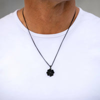 A close-up of a person wearing a black chain necklace with a black four-leaf clover pendant, styled with a white crew-neck t-shirt.