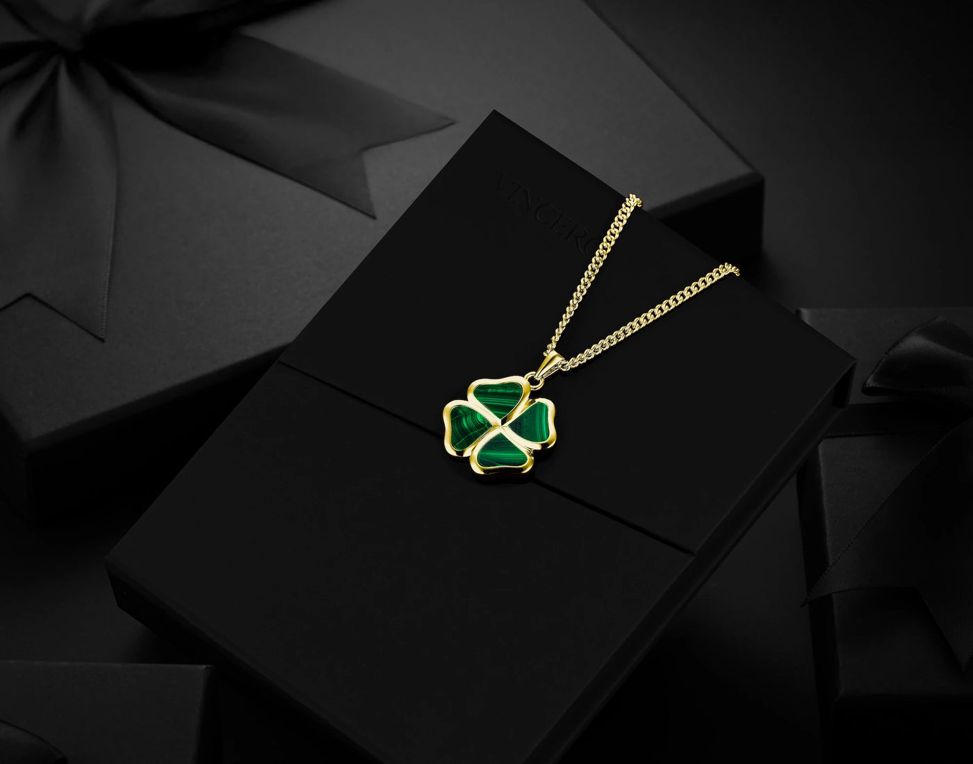 A gold chain necklace with a four-leaf clover pendant featuring green enamel inlay, displayed on a black gift box with a black ribbon bow.
