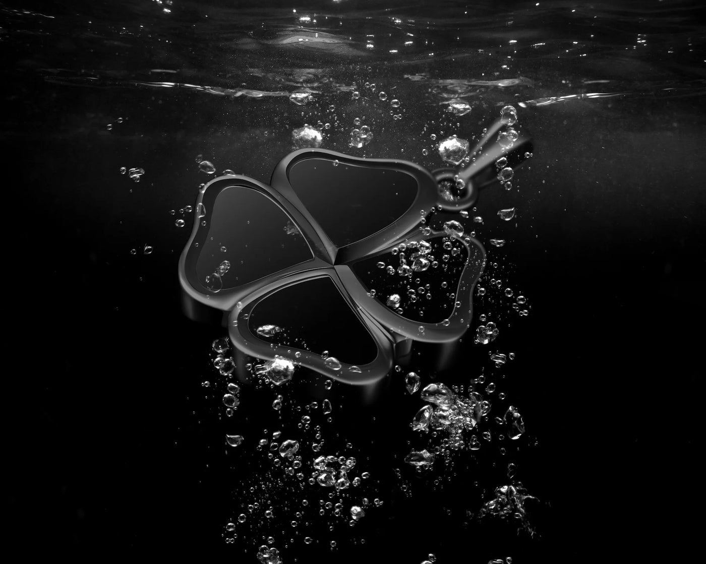 A dark metallic four-petal charm or pendant with a small loop at the top, floating underwater with bubbles around it against a black water background.
