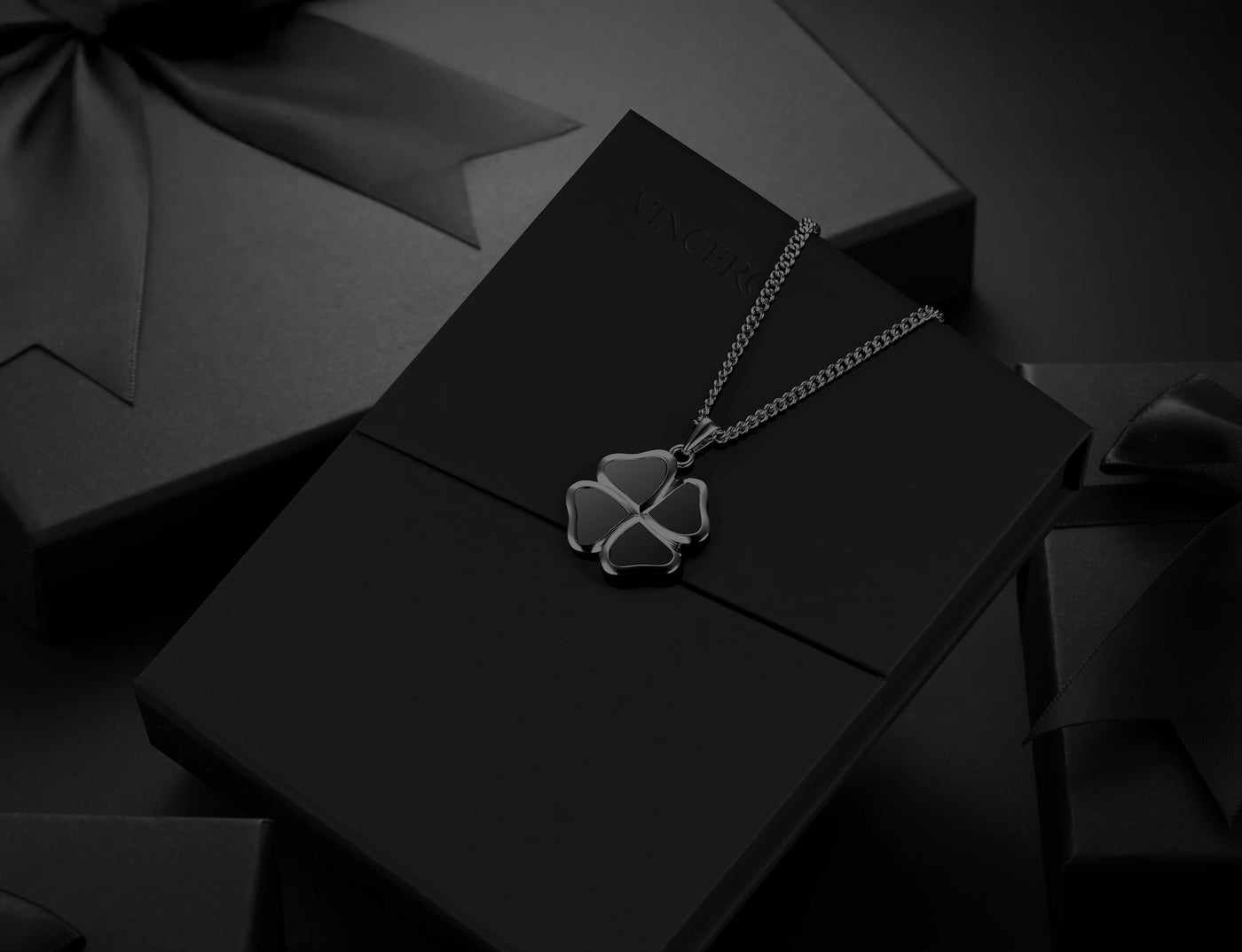 A silver-toned four-leaf clover pendant necklace resting on a black presentation box, with dark gift boxes and ribbons in the background.