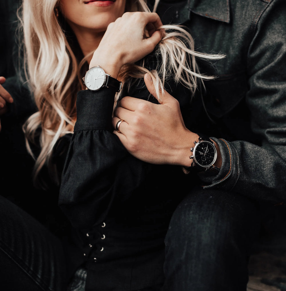 Two people in dark clothing wearing wristwatches, shown in a close cropped lifestyle shot. One watch has a white dial and black strap, and the other has a black dial and black strap.