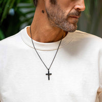Man wearing a black cross necklace against a blurred green background