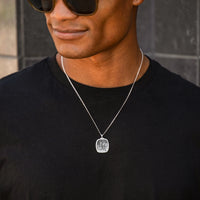 Man wearing a black shirt and silver necklace with a pendant against a gray tiled wall.