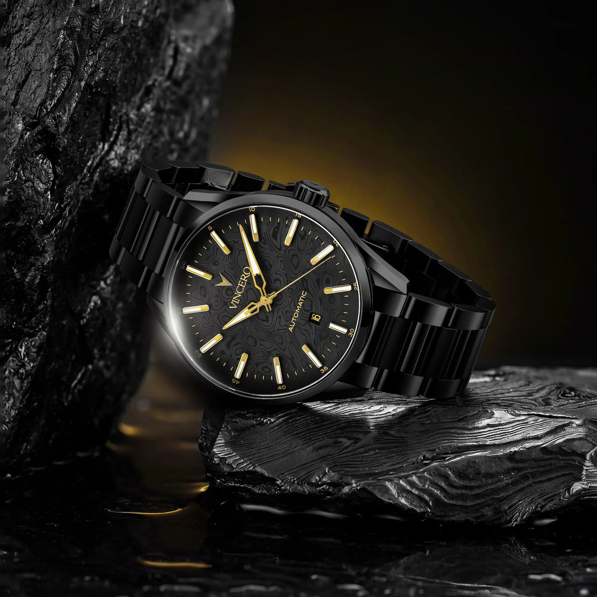A black stainless steel wristwatch with a black dial and gold-tone hands and indices, resting on dark textured rocks with a warm yellow glow in the background.