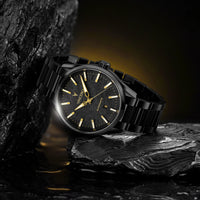 A black stainless steel wristwatch with a black dial and gold-tone hands and indices, resting on dark textured rocks with a warm yellow glow in the background.