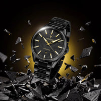 A black Seiko automatic wristwatch with a matching black metal bracelet, shown floating above shattered black glass on a dark background with a warm yellow glow behind it.