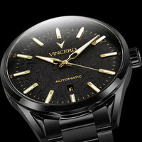 A close-up of a Vincero Automatic watch with a black dial, gold-tone hands and markers, and a black metal case and bracelet against a black background.