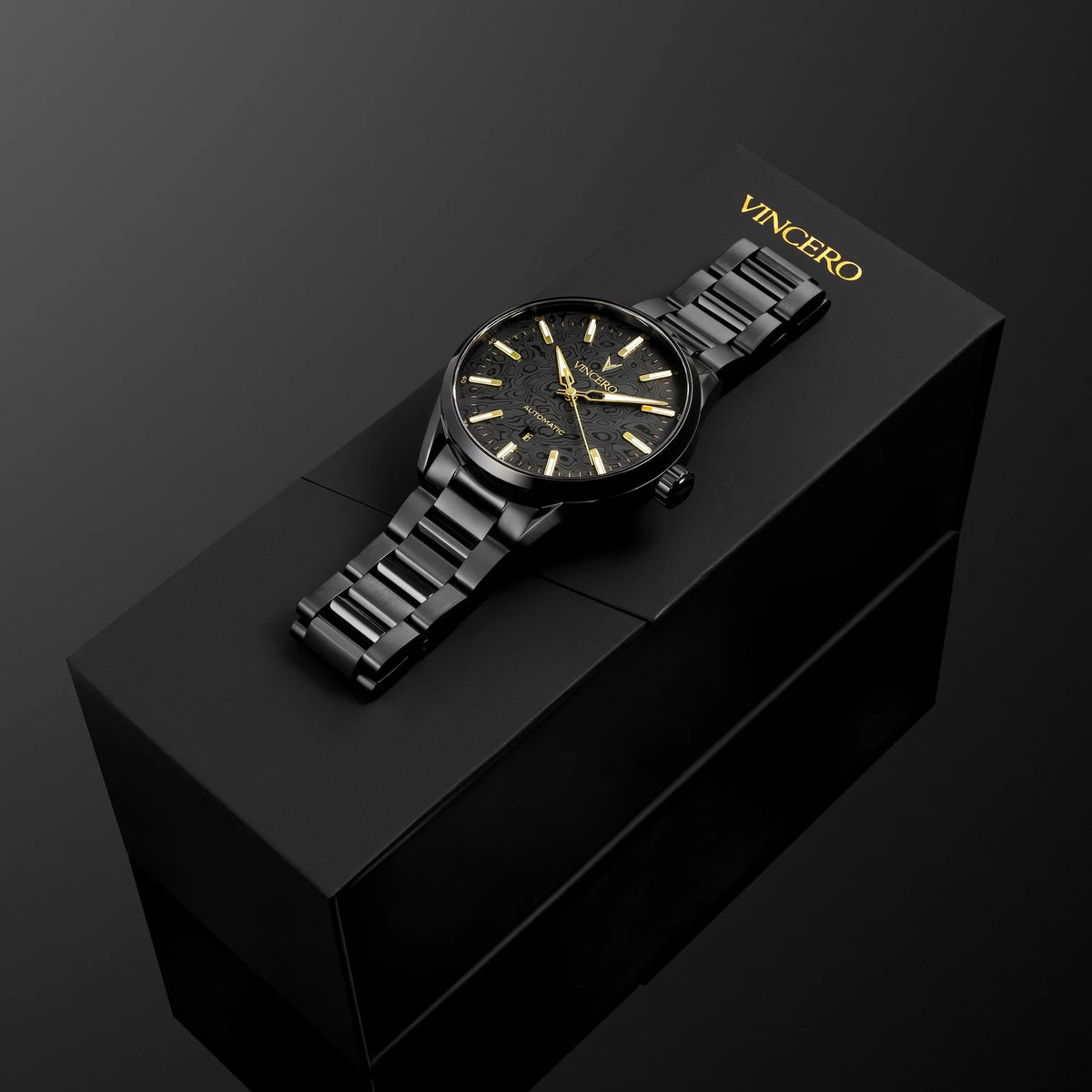 A black VINCERO wristwatch with a black metal link bracelet and gold-tone dial details, displayed diagonally on top of its black presentation box against a dark background.