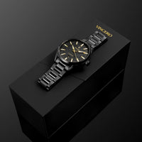 A black VINCERO wristwatch with a black metal link bracelet and gold-tone dial details, displayed diagonally on top of its black presentation box against a dark background.