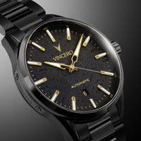 A close-up studio shot of a VINCERO automatic wristwatch with a black case, black link bracelet, and a black dial featuring gold-tone hands, markers, and text.