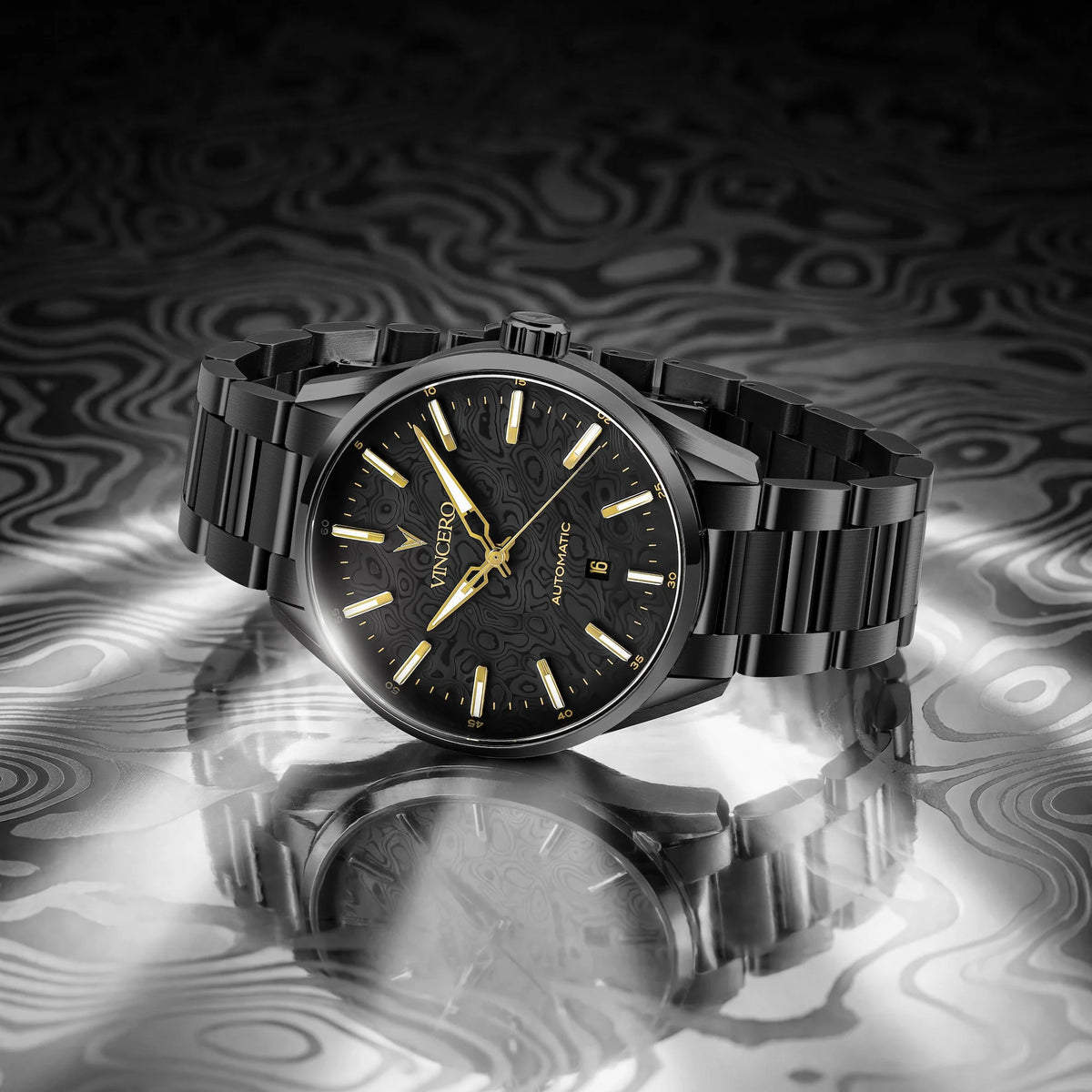 A black stainless steel wristwatch with a black dial, gold-tone indices, and a black link bracelet, displayed on a reflective surface with a dark, wavy patterned background.
