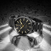 A black stainless steel wristwatch with a black dial, gold-tone indices, and a black link bracelet, displayed on a reflective surface with a dark, wavy patterned background.