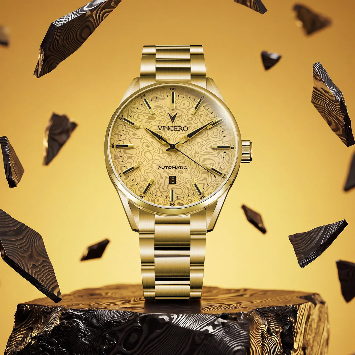 A gold-tone wristwatch with a patterned beige dial and matching metal bracelet, displayed on a wood block against a warm gold background with floating dark wood shards. The brand name VINCERO is visible on the dial.