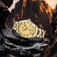 A gold-tone stainless steel wristwatch with a textured dial and matching metal bracelet, displayed on dark rocks with a mountainous outdoor background.