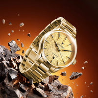 A gold-tone stainless steel wristwatch with a patterned gold dial and matching gold bracelet, shown at an angle amidst floating metallic fragments on a dark brown and orange gradient background.