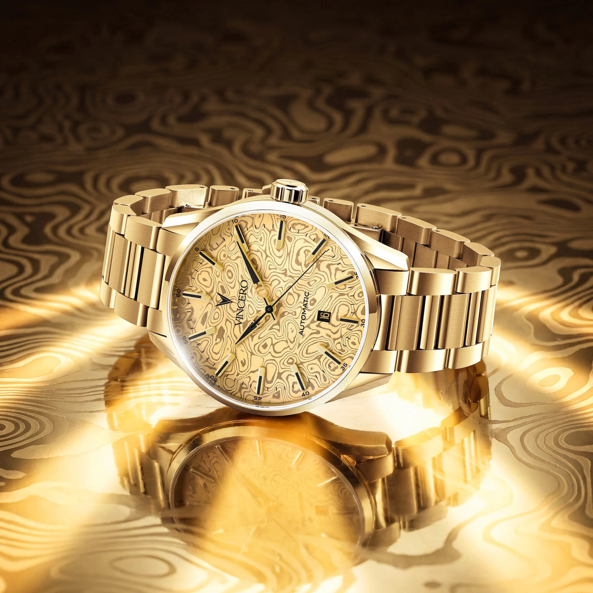 A gold-tone stainless steel wristwatch with a patterned dial and matching gold bracelet, displayed on a reflective gold surface with a dark patterned background.