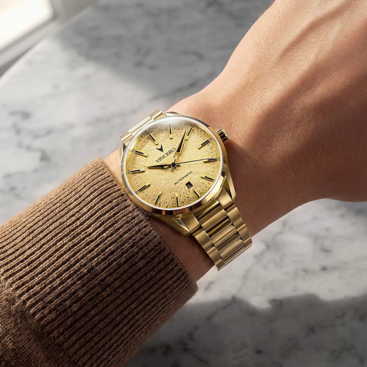 A gold-tone wristwatch with a matching gold dial and link bracelet, shown worn on a person's wrist over a brown ribbed sweater sleeve, set against a gray marble surface.