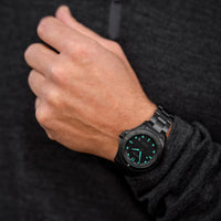 Black wristwatch with green glowing face on a person's wrist against a dark background