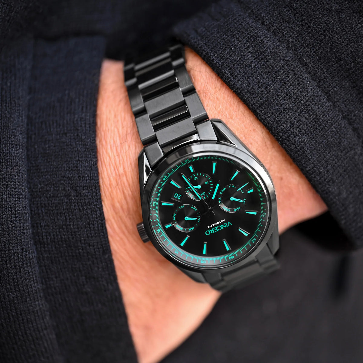 Hand wearing a black wristwatch with a dark face and green accents on a dark background