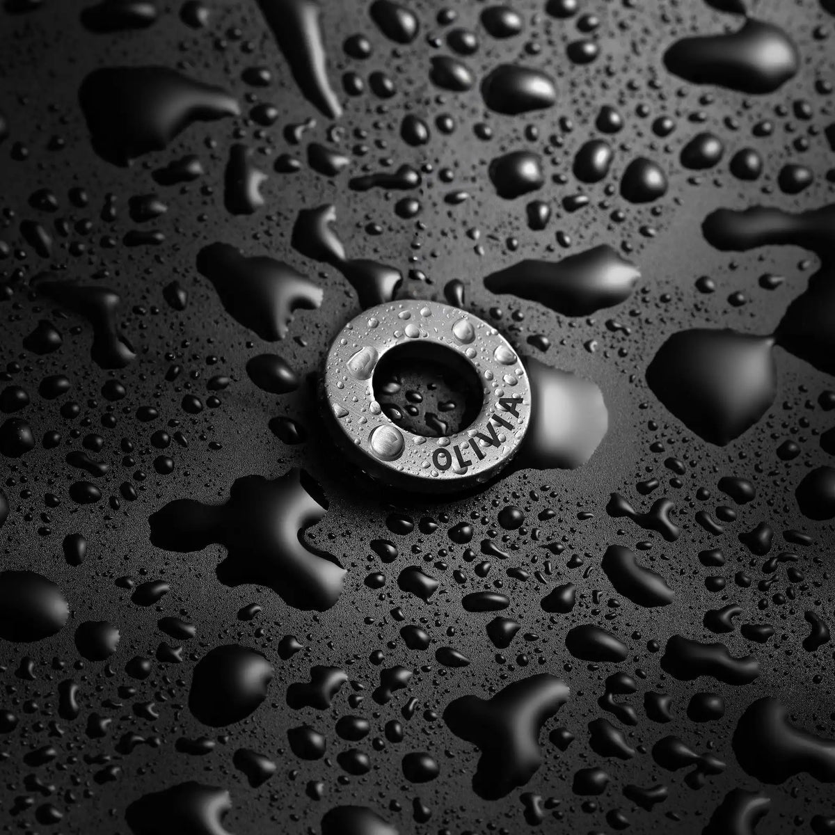 Metallic sink drain cover with 'Olivia' branding on a black surface with water droplets.