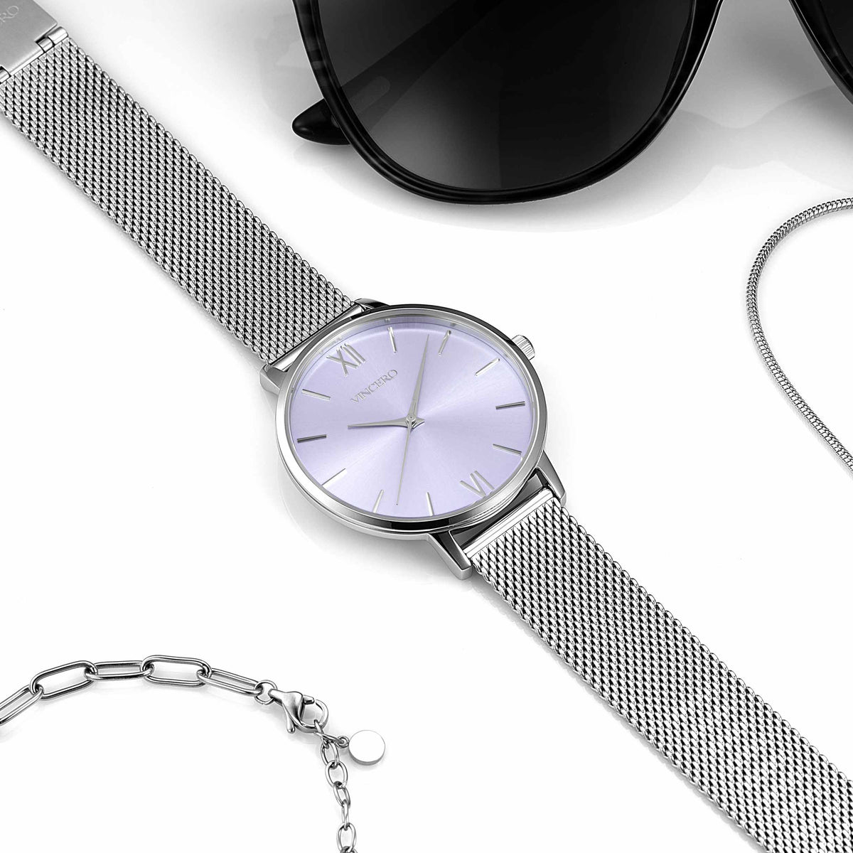 Silver watch with mesh band on a white surface with sunglasses and necklace.