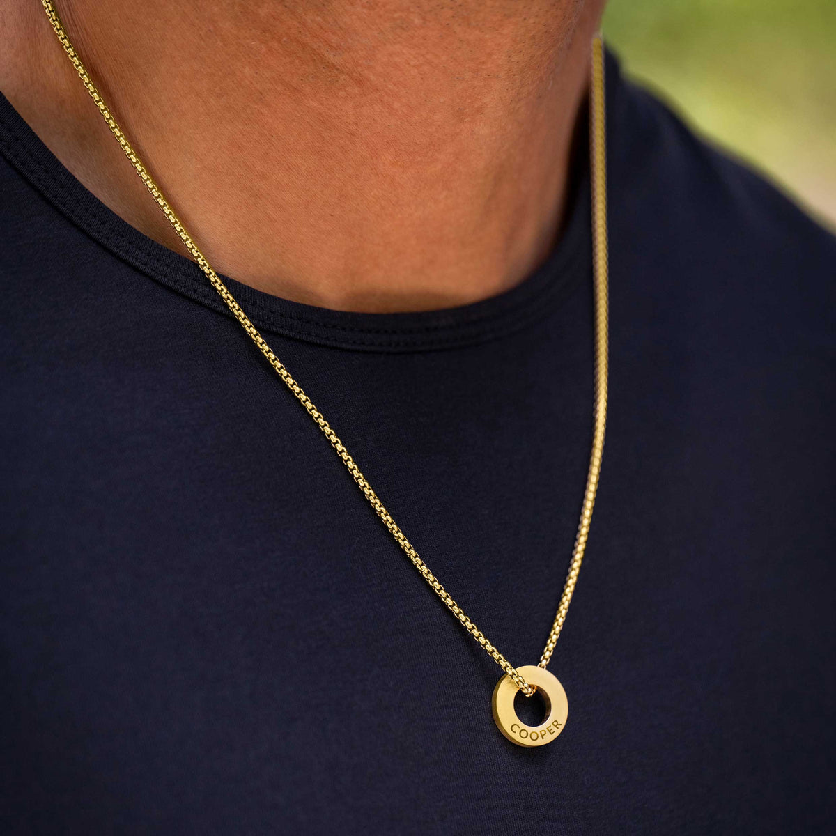A close-up of a person wearing a gold chain necklace with a circular pendant engraved with the name 'COOKS'. The person is wearing a dark navy blue t-shirt, with a softly blurred green background visible in the upper right.