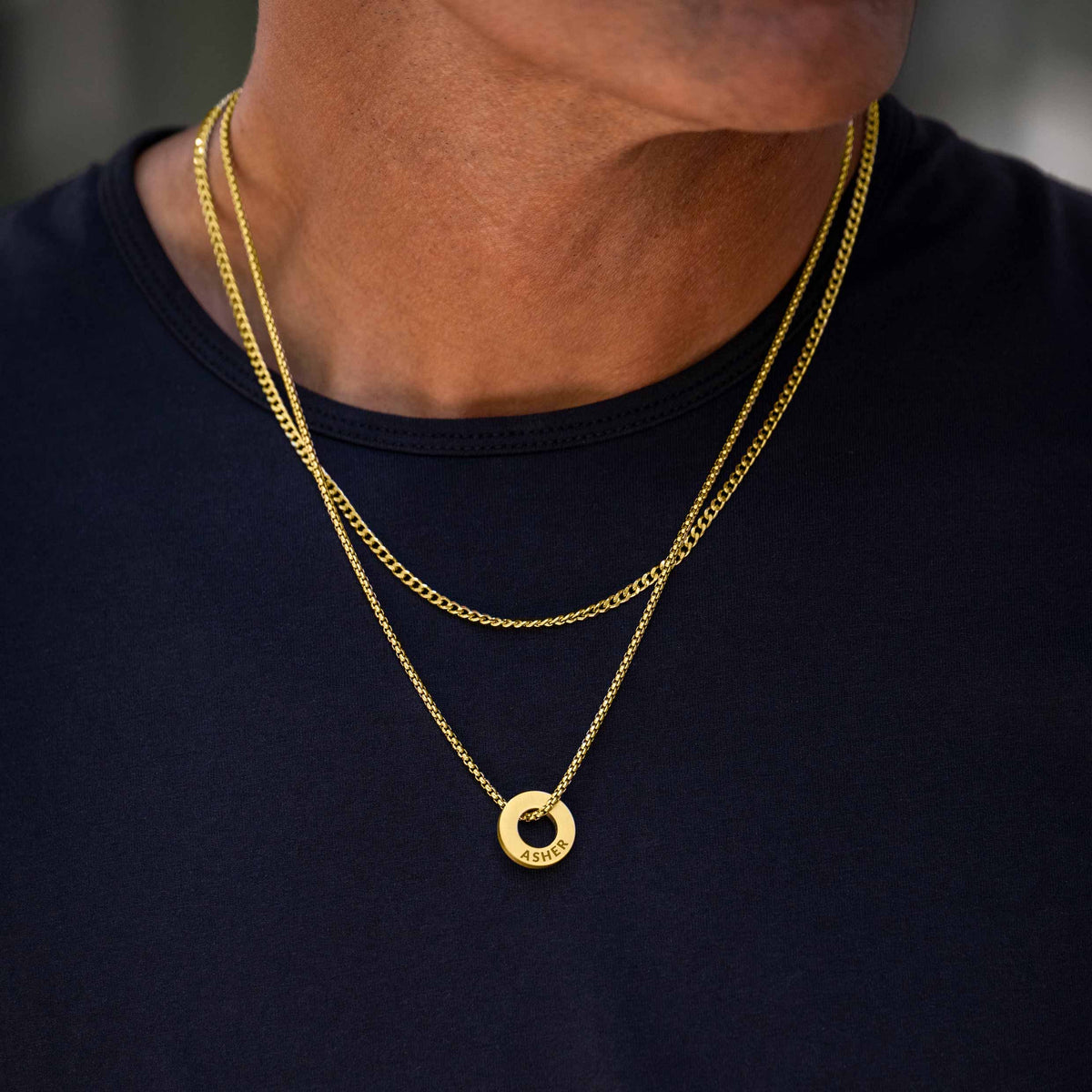 A close-up of a person wearing two layered gold necklaces, including a circular pendant necklace, over a dark navy blue t-shirt.