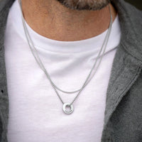 Silver necklace with a circular pendant worn by a person on a white shirt.