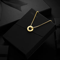 A gold-tone circular pendant necklace with the name 'VINCINO' engraved on the disc, resting on a black presentation box. The box features the brand name 'VINCINO' debossed on the top, and the necklace is displayed against a dark, moody background.