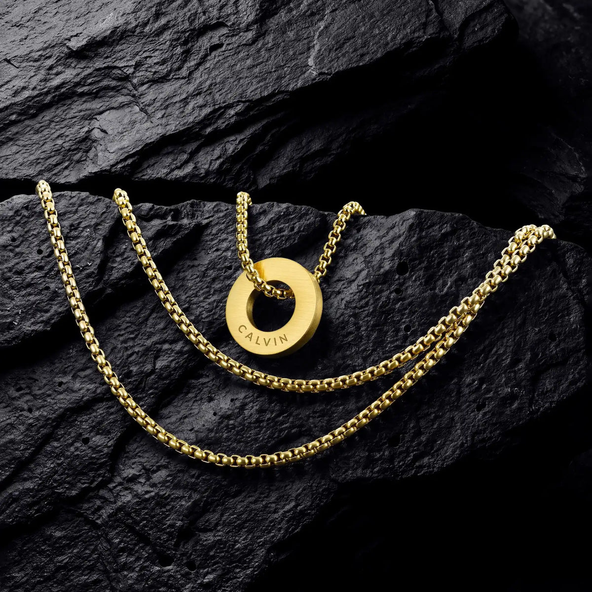 A gold chain necklace with a circular ring pendant engraved with the brand name 'CALVIN', displayed on a dark, textured rock surface.