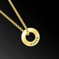 A gold chain necklace with a circular ring pendant engraved with the name 'ASHER', shown against a solid black background.