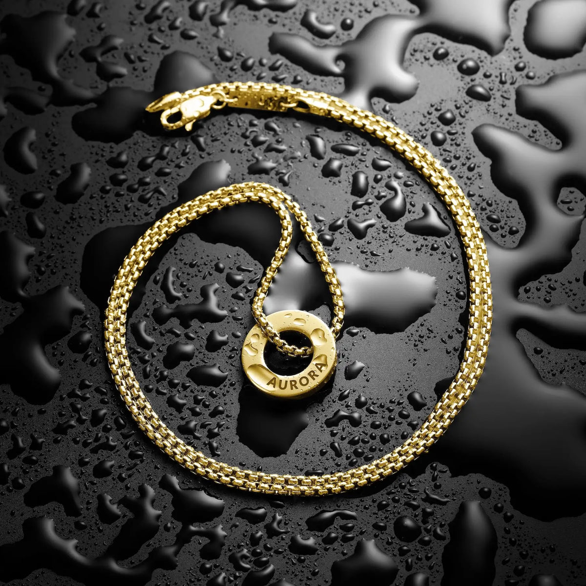 A gold chain necklace with a circular ring pendant engraved with the brand name 'AURORA', arranged on a glossy black surface covered in water droplets.