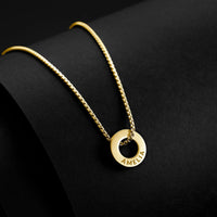 A gold necklace with a textured chain and a circular pendant engraved with the name 'AMELIA', displayed on a black curved surface.