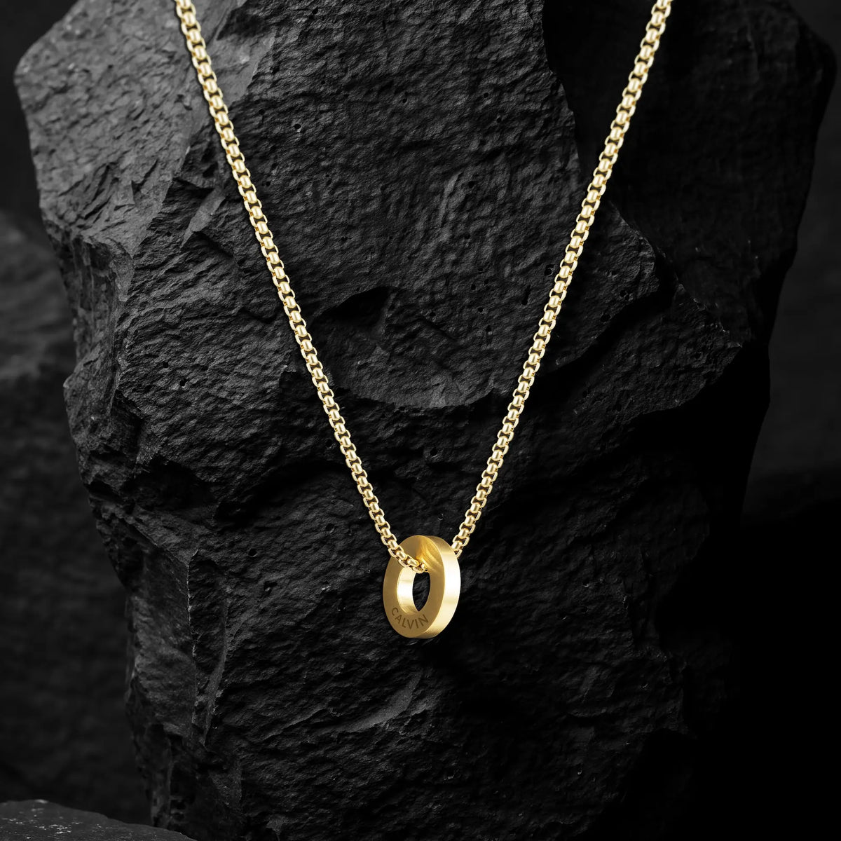A gold chain necklace with a polished interlocking double-ring pendant, displayed on a textured black rock against a dark background.