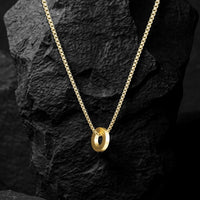 A gold chain necklace with a polished interlocking double-ring pendant, displayed on a textured black rock against a dark background.