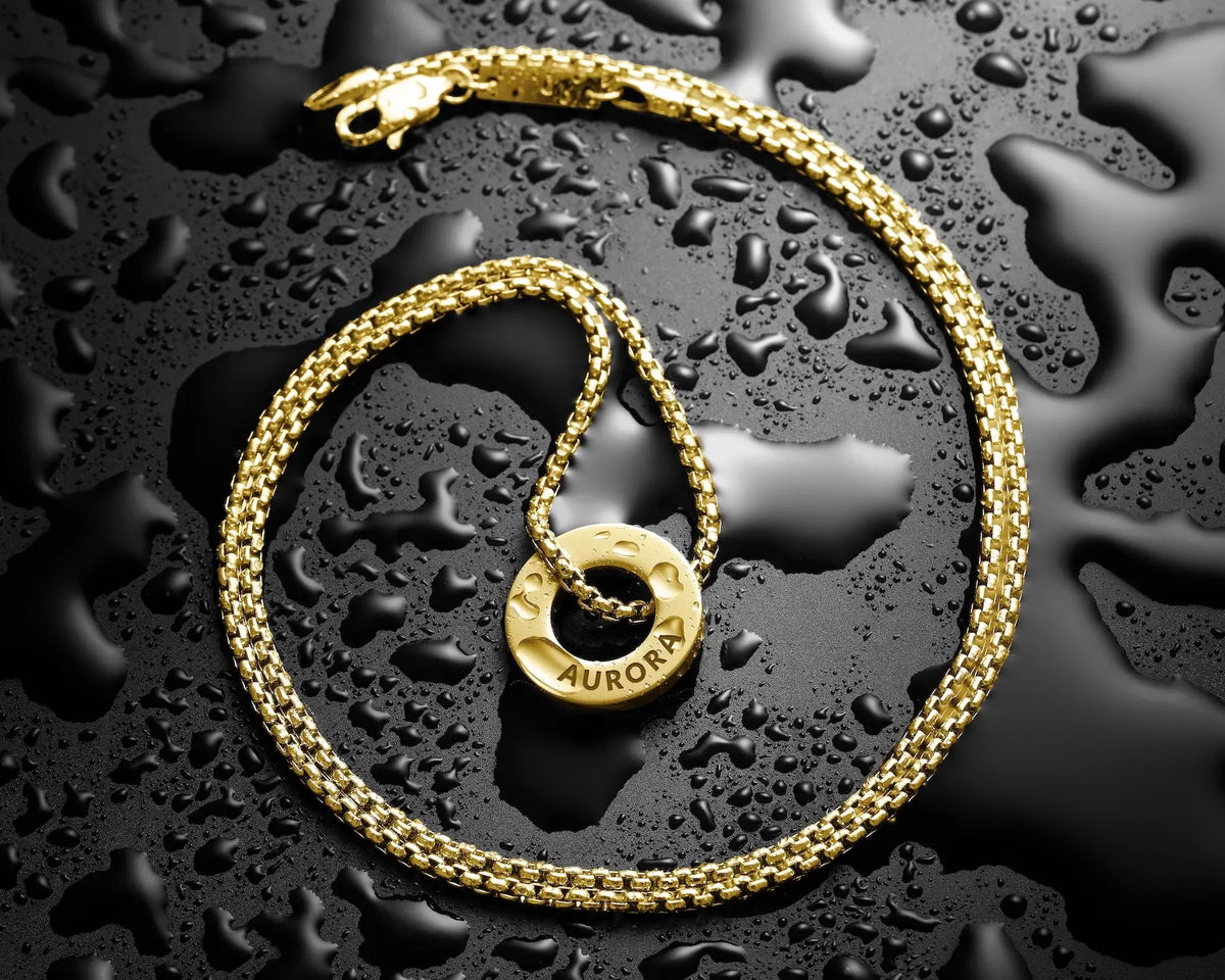 A gold chain necklace with a circular ring pendant engraved with the name 'AURORA', arranged on a glossy black surface covered in water droplets.