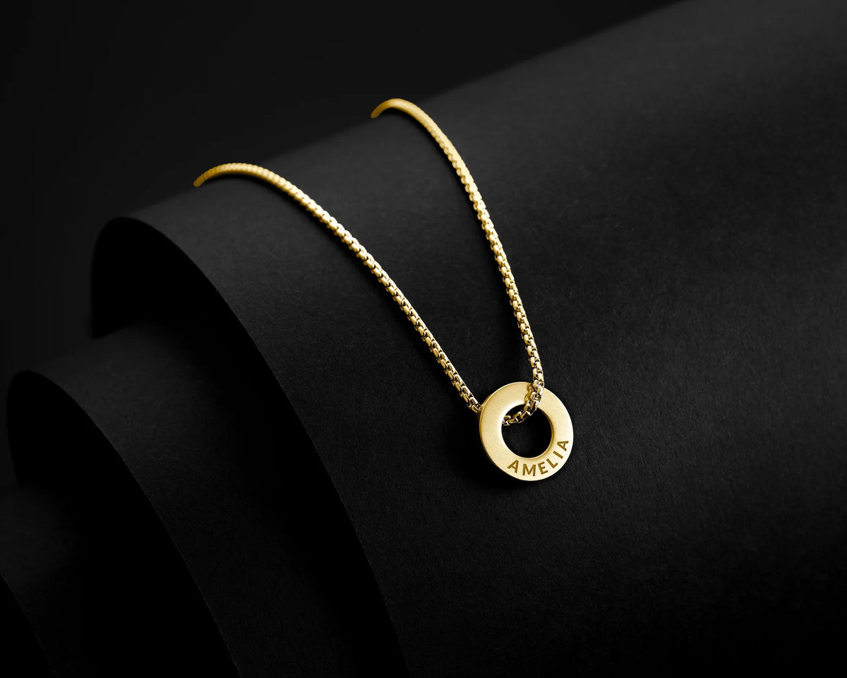 A gold necklace with a textured chain and a circular pendant engraved with the word 'AMBIT', displayed on a black curved stand against a dark background.