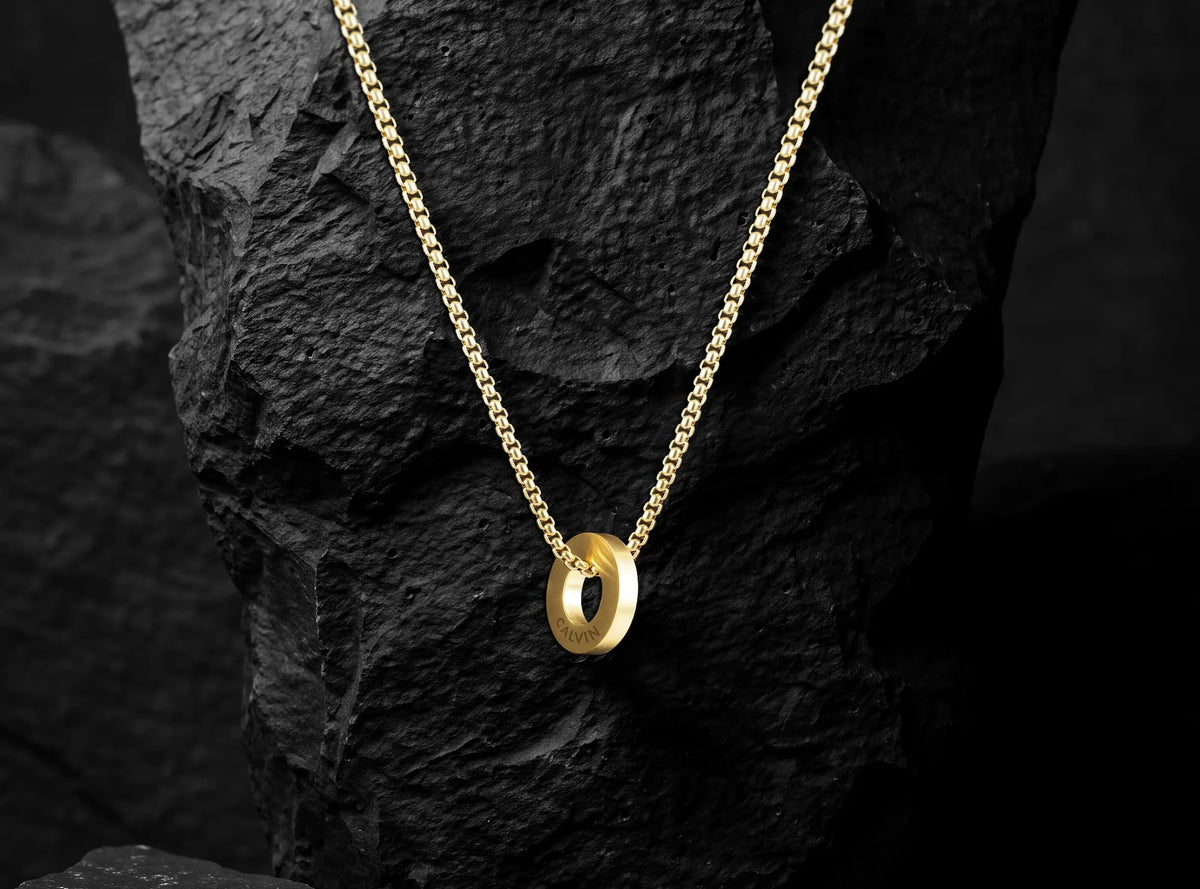 A gold chain necklace with a polished oval pendant, displayed against a dark textured rock background.