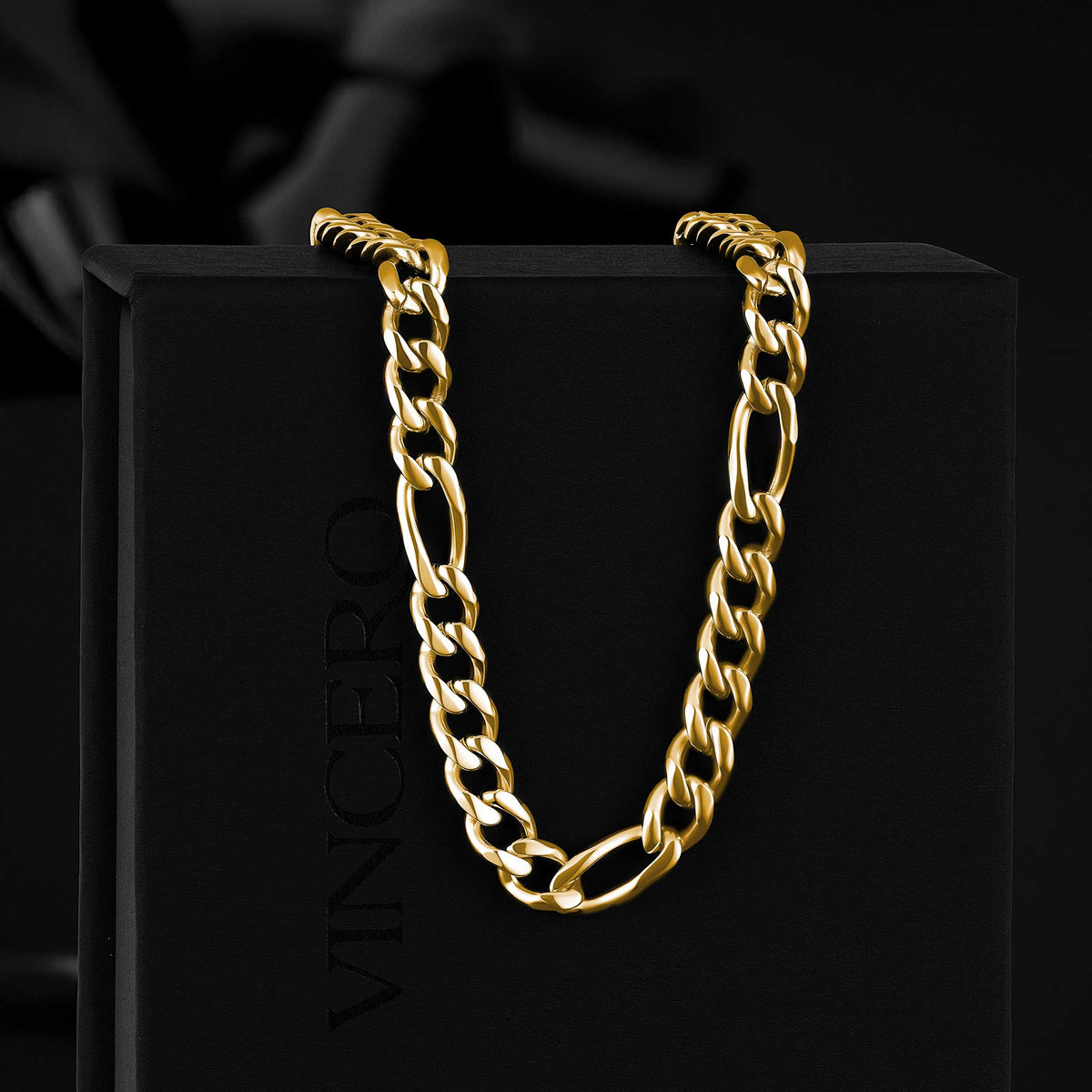 A gold-tone Figaro chain necklace draped over a black display box with the brand name VINCERO partially visible, set against a dark black background.