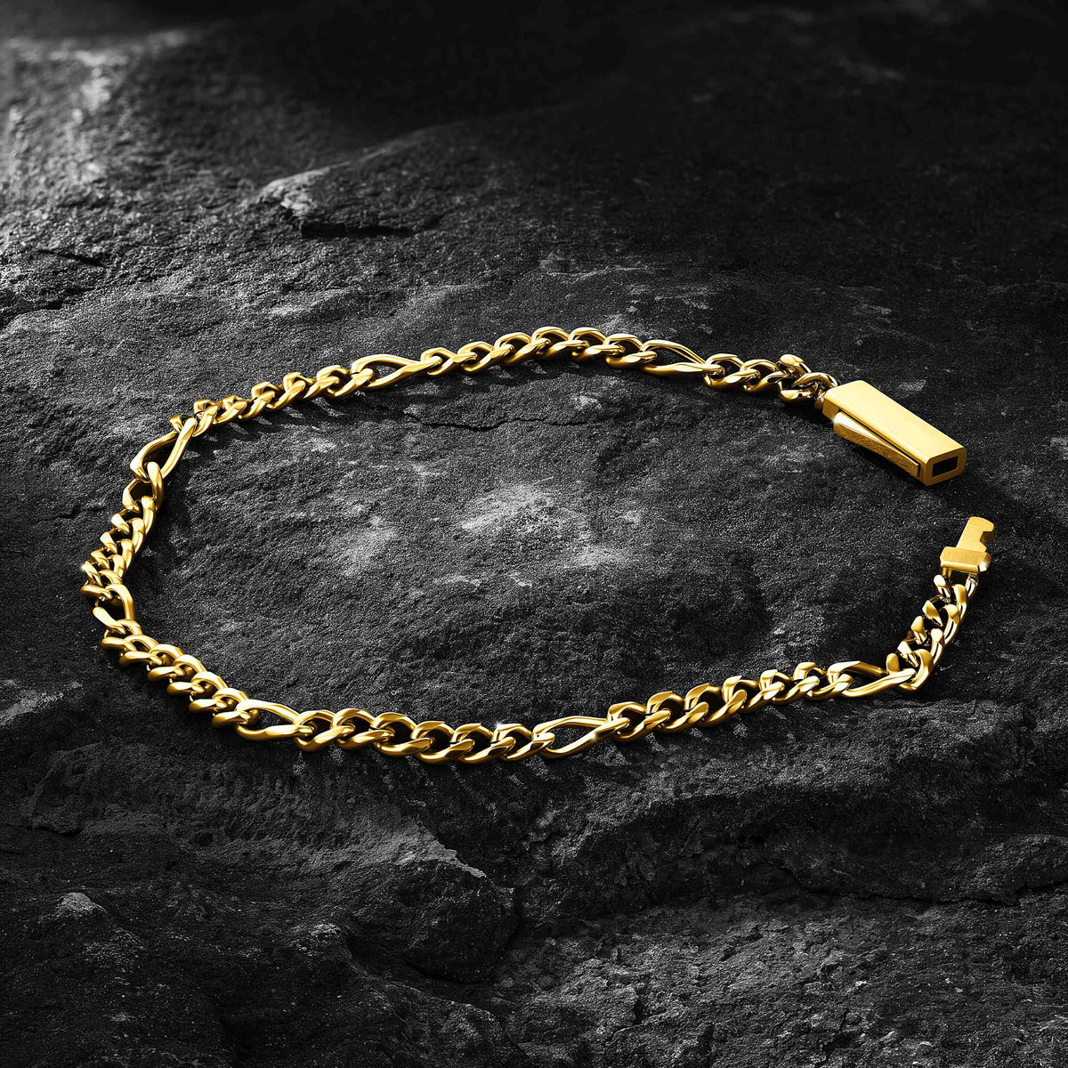A gold-toned figaro link chain bracelet with a rectangular clasp, arranged in an oval on a dark textured rock surface.