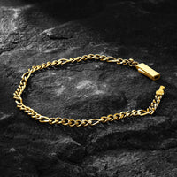 A gold-toned figaro link chain bracelet with a rectangular clasp, arranged in an oval on a dark textured rock surface.