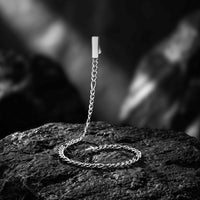 A silver chain accessory with a rectangular metal clasp and a braided chain section, displayed on a dark textured rock against a blurred black-and-white natural background.