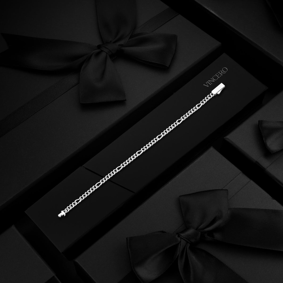 A silver-toned chain bracelet displayed on a black gift box with a black ribbon bow, surrounded by matching black boxes. The brand name VINCI GO is printed on the box.