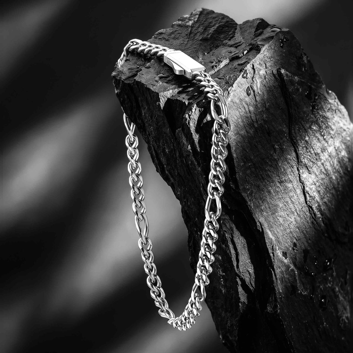 A silver-toned metal chain necklace draped over a dark textured rock, shown in a high-contrast black and white photograph.