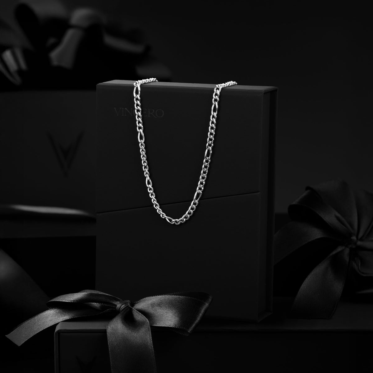 A silver-toned chain necklace draped over a black presentation box, with matching black gift boxes and satin bows in a dark studio setting.