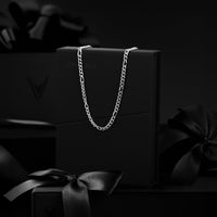 A silver-toned chain necklace draped over a black presentation box, with matching black gift boxes and satin bows in a dark studio setting.